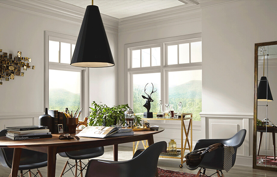 An office with large windows in the corner and a tall mirror leaning against the walls, which are painted a light neutral Agreeable Gray SW 7029. There is an oval wood table with four chairs around it with a very large black conical pendant light overhead.