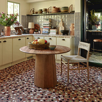 A retro-styled kitchen with cabinets and walls painted a cream color with yellow tones. The walls have open shelves and a metallic backsplash. A round pedestal wood table with one woven chair sits in the middle. The floor is a mosaic of black, white, and gray tile pieces with dark red grout. 