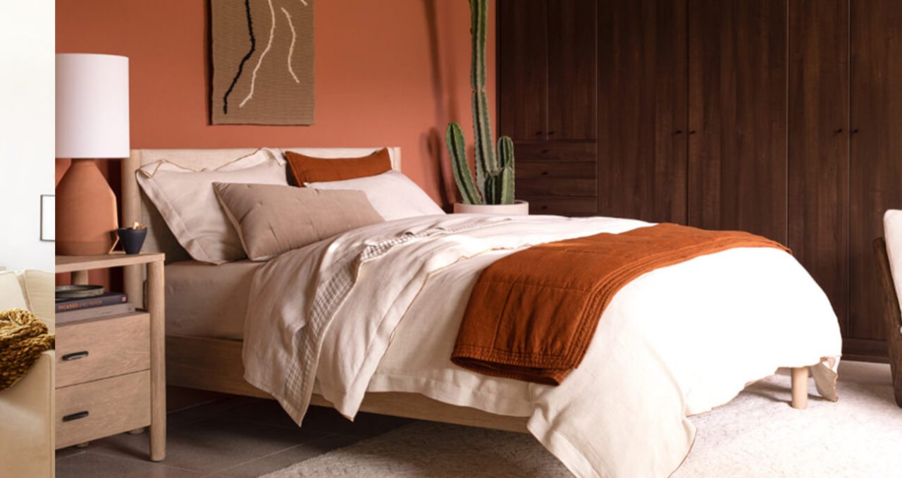A bed with neutral bedding and orange accents sits against a warm orange wall. A potted cactus sits to the right and the right wall is wood plank paneled. A nightstand with a terracotta-colored lamp sits to the left. 