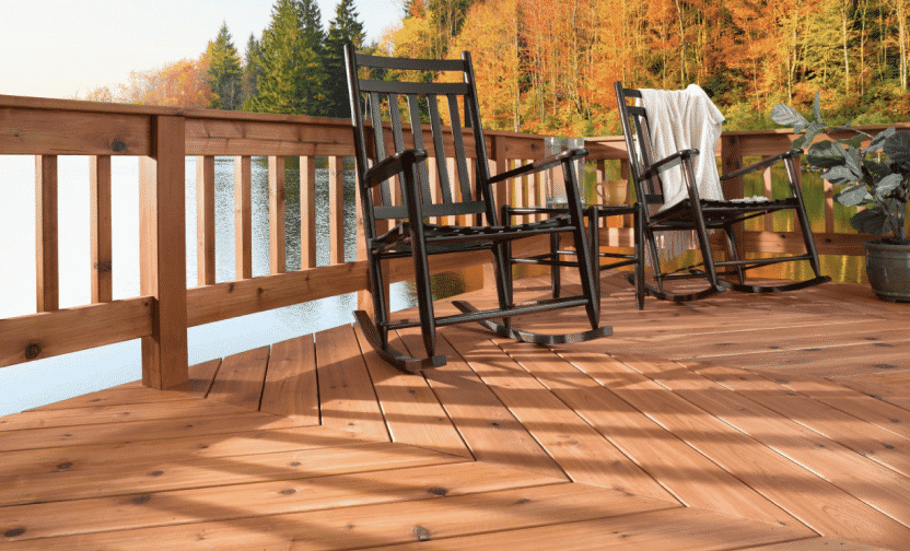A cedar colored deck on a lake with two black rocking chairs and fall leaves behind.