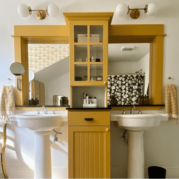 Two white pedestal sinks with a dark yellow painted cabinet in between and framed mirror painted the same by @ciarakenaston.